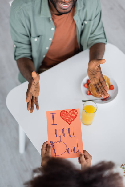 Вид сверху на африканского ребенка, держащего открытку с поздравлением "Я люблю тебя, папа, рядом с веселым мужчиной в день отца"  - Фото, изображение