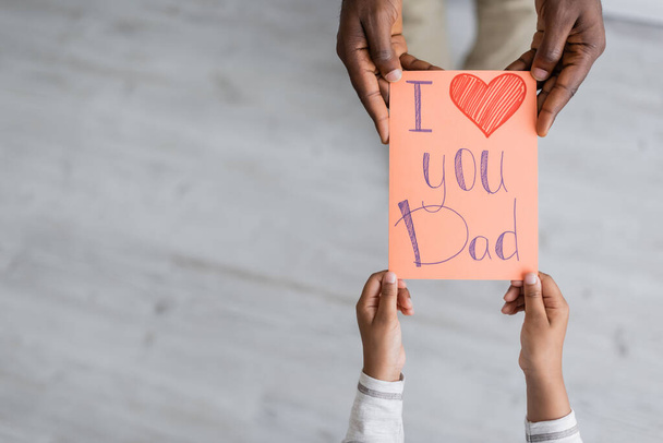 vista superior del niño afroamericano recortado sosteniendo la tarjeta de felicitación con te amo papá letras cerca del hombre en el día de los padres  - Foto, imagen