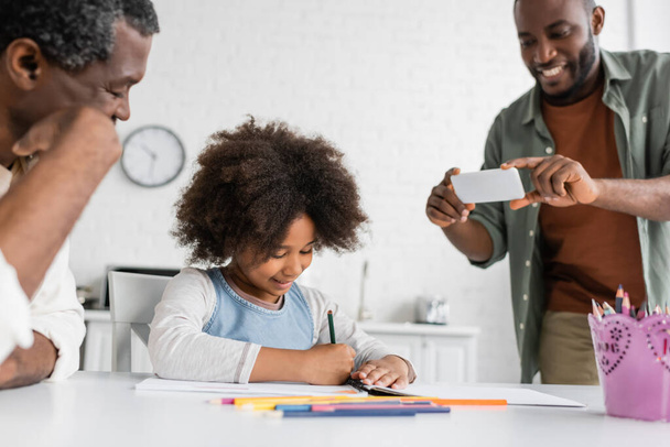 happy african american man taking photo on smartphone while his daughter and father drawing together  - Valokuva, kuva