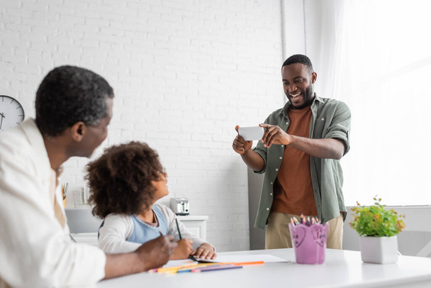 fröhlicher afrikanisch-amerikanischer Mann beim Fotografieren auf dem Smartphone, während Tochter und Vater zusammen zeichnen  - Foto, Bild