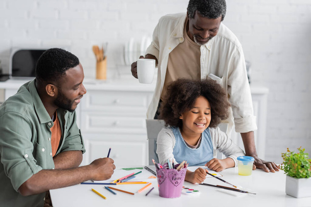 Positive african american man sitting near daughter painting and father holding cup of coffee at home  - Foto, Imagen