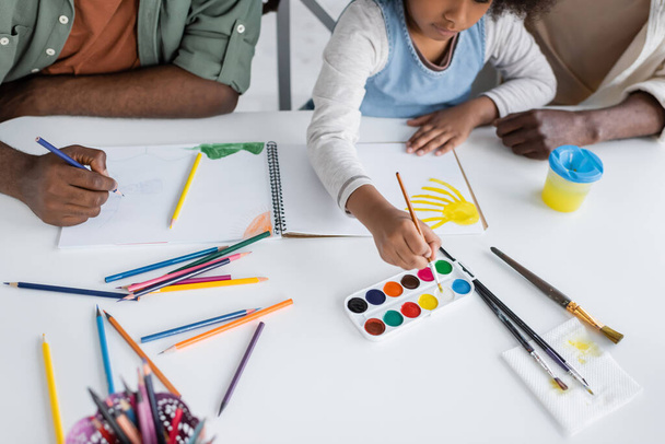 Vista recortada de niños afroamericanos y padres pintando en cuaderno de bocetos en casa  - Foto, imagen