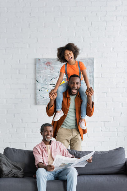 Heureux homme et enfant afro-américain s'amuser près de grand-parent avec journal sur le canapé à la maison  - Photo, image