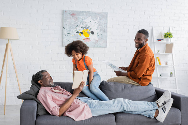 笑顔アフリカ系アメリカ人の女の子座っています近くおじいちゃんと本とお父さんとともに家に  - 写真・画像