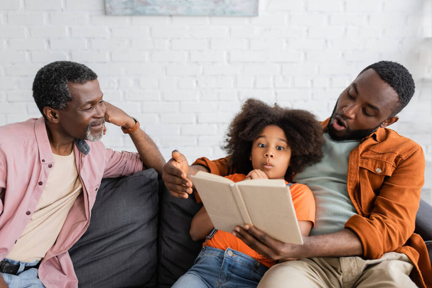 Shocked african american father and daughter reading book near grandparent at home  - Photo, Image