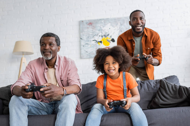 KYIV, UKRAINE - JULY 17, 2021: Cheerful african american kid playing video game with father and granddad at home  - Photo, image