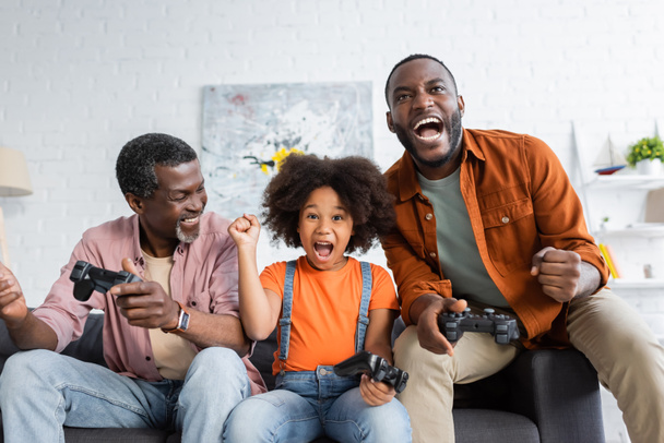 KYIV, UKRAINE - JULY 17, 2021: Excited african american girl and dad holding joysticks near grandfather at home  - Foto, Imagen