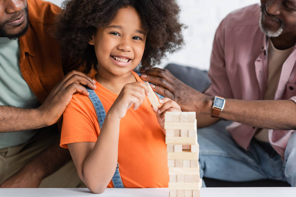 Heureux gamin afro-américain regardant la caméra près des parents et des blocs de bois jeu à la maison  - Photo, image