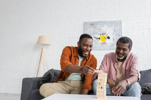 Alegre hombre afroamericano jugando bloques de madera juego con el padre maduro en la sala de estar  - Foto, Imagen