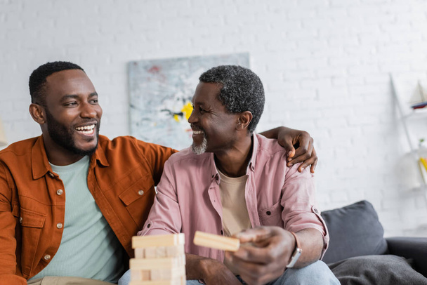 African american man hugging mature father near blurred wood blocks game at home  - 写真・画像