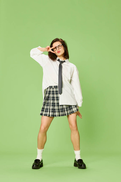 Cheerful funny mood. Full-length portrait of young beautiful woman in checkered skirt and white shirt posing against green studio background. Concept of youth, fashion, emotions, facial expression. Ad - Photo, Image