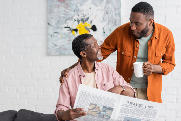 Afrikai amerikai férfi kezében csésze kávét, és beszél apával újság otthon  - Fotó, kép
