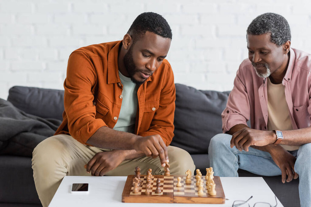 Αφροαμερικάνος παίζει σκάκι με τον ώριμο πατέρα του στο σαλόνι. - Φωτογραφία, εικόνα