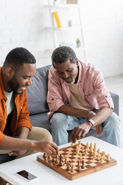 Afro-américain jouer aux échecs avec un fils souriant près d'un smartphone à la maison  - Photo, image
