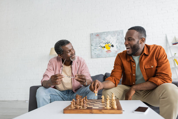 Overjoyed african american man playing chess with mature dad near smartphone with blank screen at home  - 写真・画像