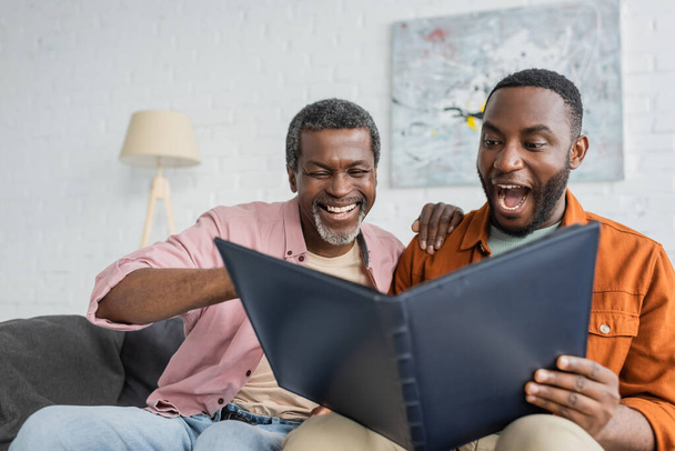 Cheerful and excited african american father and son looking at photo album at home  - Foto, afbeelding