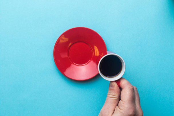 An Espresso cup of black coffee being held by a hand on a blue background with a saucer - Photo, Image