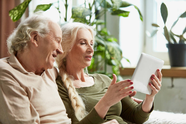 Happy positive senior couple, retired man and woman sitting on sofa at home and having online video call on tablet. Grandparents. Communication. Concept of family, relationship, modern technologies - Photo, Image