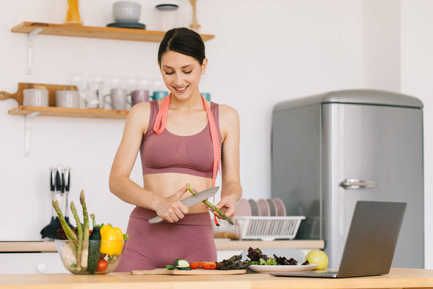 Mutlu spor kadın blogcu portresi kuşkonmaz pişiriyor ve mutfaktaki dizüstü bilgisayarda sağlıklı beslenme konulu video konferansı düzenliyor. - Fotoğraf, Görsel