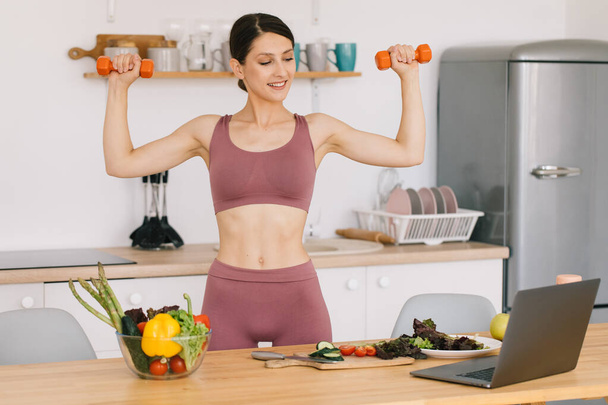 Portrait of happy sporty woman blogger showing biceps with dumbbells and leading video conference on healthy eating on laptop in kitchen - Photo, Image