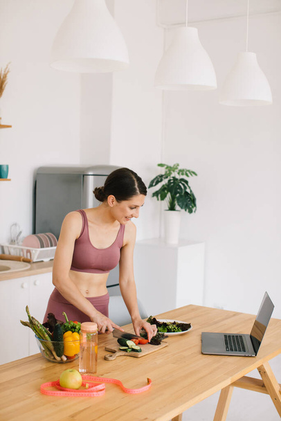 Спортивная женщина-блоггер диетолог готовит салат со свежими овощами и проводит видеоконференцию по здоровому питанию на ноутбуке на кухне - Фото, изображение