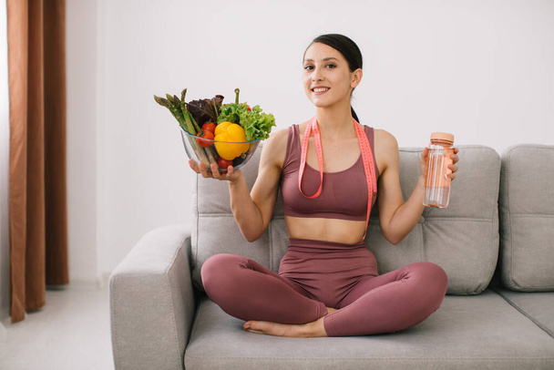 Молодая спортсменка сидит на диване, держа в руках разнообразные овощи, бутылку воды и завернутую линейку, концепцию потери веса и здорового питания. - Фото, изображение