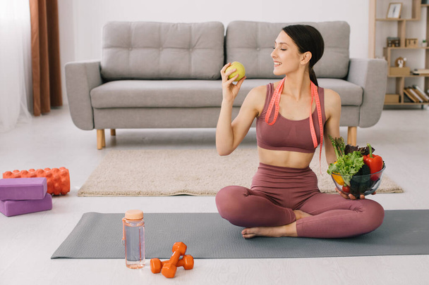 Young sporty woman sitting on yoga mat and holding variety of vegetables and apple, concept of weight loss and healthy eating. - Foto, imagen