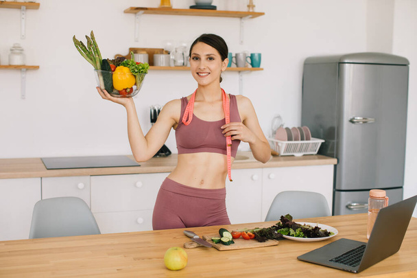 Portrait of happy sporty woman blogger holding plate of fresh vegetables and leading video conference on healthy eating on laptop in kitchen - Zdjęcie, obraz