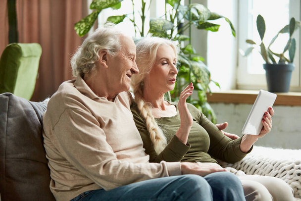 Happy cheerful senior couple, retired man and woman sitting on sofa at home and talking on online video call via tablet. Grandparents. Concept of family, relationship, modern technologies, love. - Foto, Bild