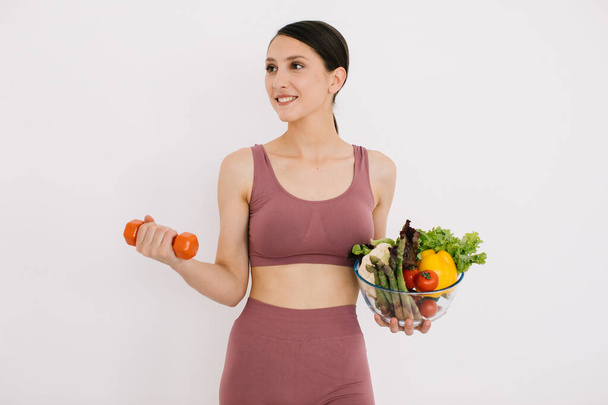 様々な健康的な野菜のトレイを持つ美しい幸せな若い女性と白い背景に隔離されたダンベルで彼女の筋肉を示す - 写真・画像