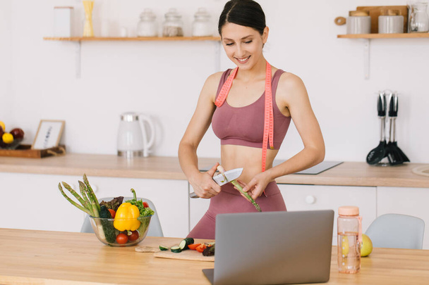 Retrato de blogueiro mulher desportiva feliz fazendo espargos cozinhar e vídeo conferência líder sobre o tema da alimentação saudável no laptop na cozinha - Foto, Imagem