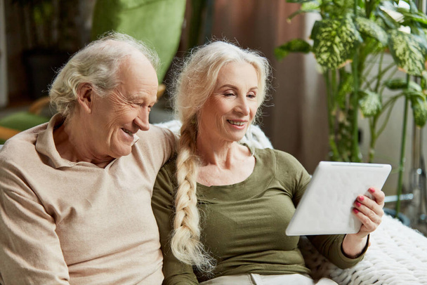 Happy smiling people, retired couple, man and woman sitting on couch at home and looking on tablet. Watching movies, talking with relatives online. Concept of family, relationship, modern technologies - Foto, Bild
