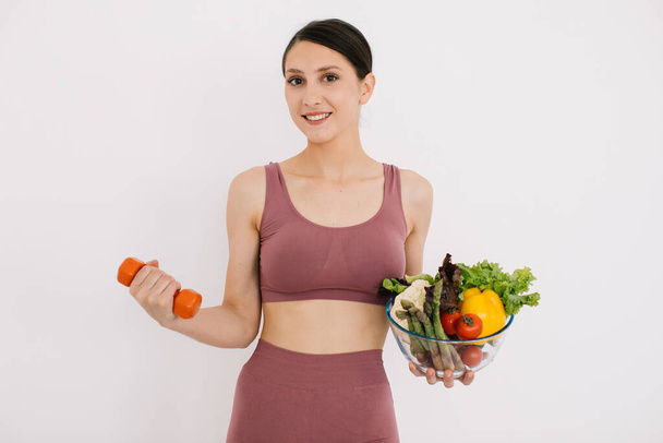 Piękna szczęśliwa młoda kobieta z tacą różnych zdrowych warzyw i pokazując swoje mięśnie z hantlami izolowane na białym tle - Zdjęcie, obraz