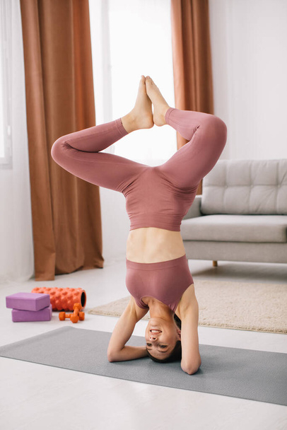 Attractive young woman stands on her head and upside down and doing fitness exercises at home. - Zdjęcie, obraz