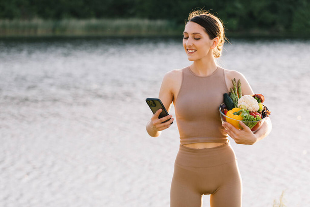 Happy sporty woman looking at phone and holding fresh vegetables outdoors - Photo, Image