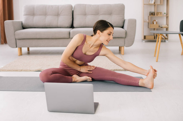 Sportowa młoda kobieta ćwiczy jogę przed laptopem, ogląda samouczki online. - Zdjęcie, obraz
