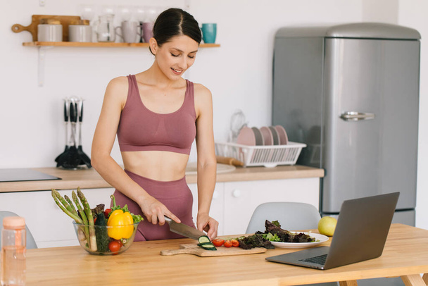 Szczęśliwa kobieta sportowiec blogger dietetyk przygotować sałatkę ze świeżych warzyw i prowadzi wideokonferencję na temat zdrowego odżywiania się na laptopie w kuchni - Zdjęcie, obraz