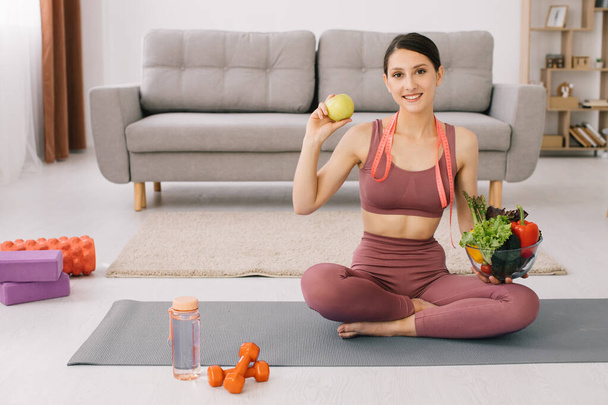Nuori urheilullinen nainen istuu joogamatolla ja pitää erilaisia vihanneksia ja omenoita, laihtuminen ja terveellinen syöminen. - Valokuva, kuva