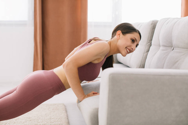 Jeune femme attrayante et en bonne santé faisant push-ups sur le canapé tout en se reposant à la maison - Photo, image