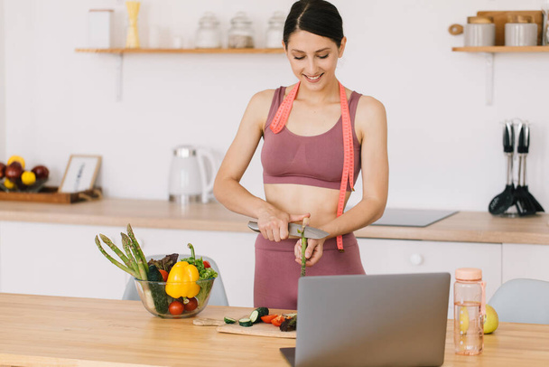 Portrait of happy sporty woman blogger making asparagus cooking and leading video conference on healthy eating topic on laptop in kitchen - Fotoğraf, Görsel