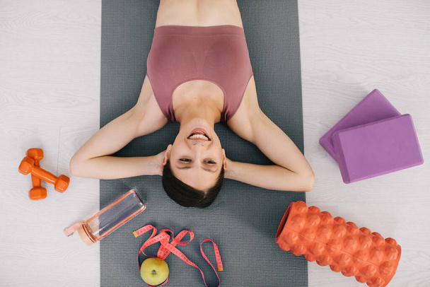 Atrakcyjna sportowa młoda kobieta leżąca na macie do jogi i wokół swojego sprzętu sportowego i jabłka, widok z góry - Zdjęcie, obraz