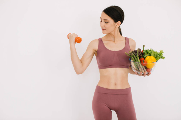Красива щаслива молода жінка з лотком різних здорових овочів і показуючи свої м'язи гантелі ізольовані на білому тлі
 - Фото, зображення