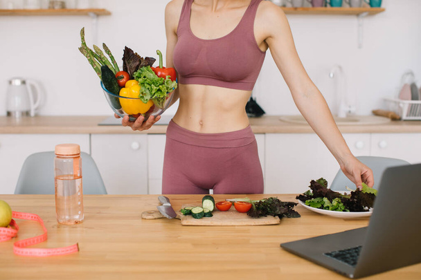 Αθλητική γυναίκα blogger διατροφολόγος προετοιμάσει μια σαλάτα με φρέσκα λαχανικά και διεξάγει ένα βίντεο συνέδριο για την υγιεινή διατροφή στο φορητό υπολογιστή στην κουζίνα - Φωτογραφία, εικόνα