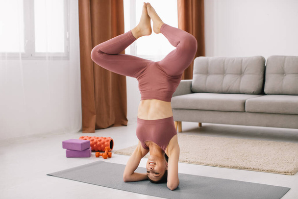 Attractive young woman stands on her head and upside down and doing fitness exercises at home. - Photo, Image