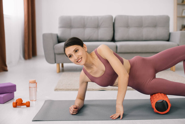 Atractiva joven mujer deportiva haciendo ejercicios en el abdomen con un rodillo en casa. - Foto, Imagen