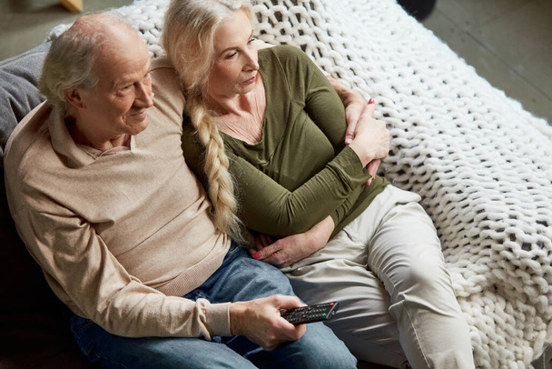 Top view image of middle-aged, mature couple, man and woman sitting on couch at home and watching tv together. Leisure time. Concept of family, relationship, retirement, lifestyle, happiness - Photo, image