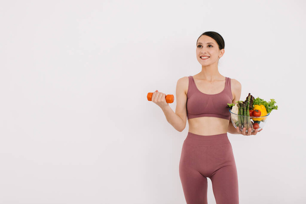 Красивая счастливая молодая женщина с подносом различных здоровых овощей и показывая свои мышцы с гантели изолированы на белом фоне - Фото, изображение