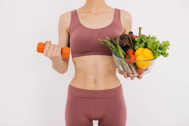 Красивая счастливая молодая женщина с подносом различных здоровых овощей и показывая свои мышцы с гантели изолированы на белом фоне - Фото, изображение
