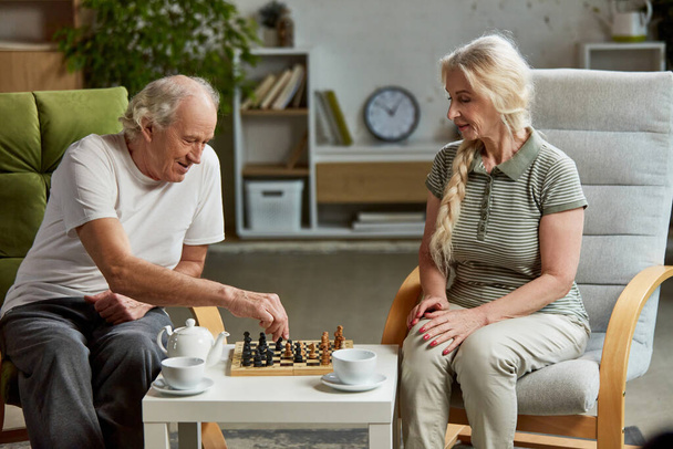 Happy elderly couple, retired man and woman spending time at home, playing chess together, drinking tea. Leisure time and hobby. Concept of family, relationship, retirement, lifestyle, happiness - Photo, image