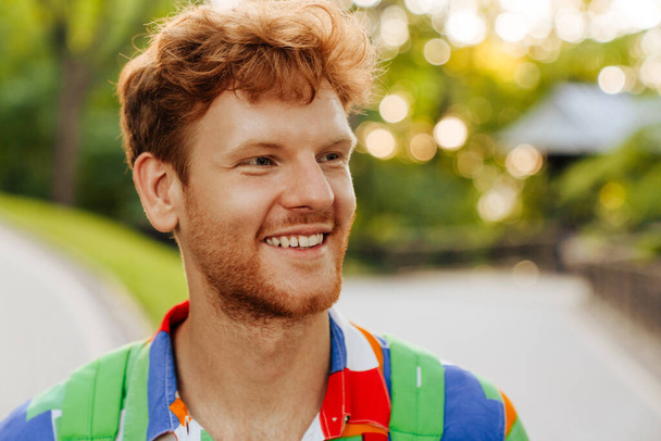 Retrato de jovem bonito sorrindo homem feliz em camisa colorida olhando para o lado, enquanto em pé no parque - Foto, Imagem